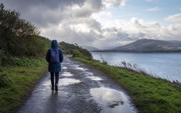 Person in a raincoat walking calmly along a quiet path after rainfall, carrying a small backpack beneath a clearing sky