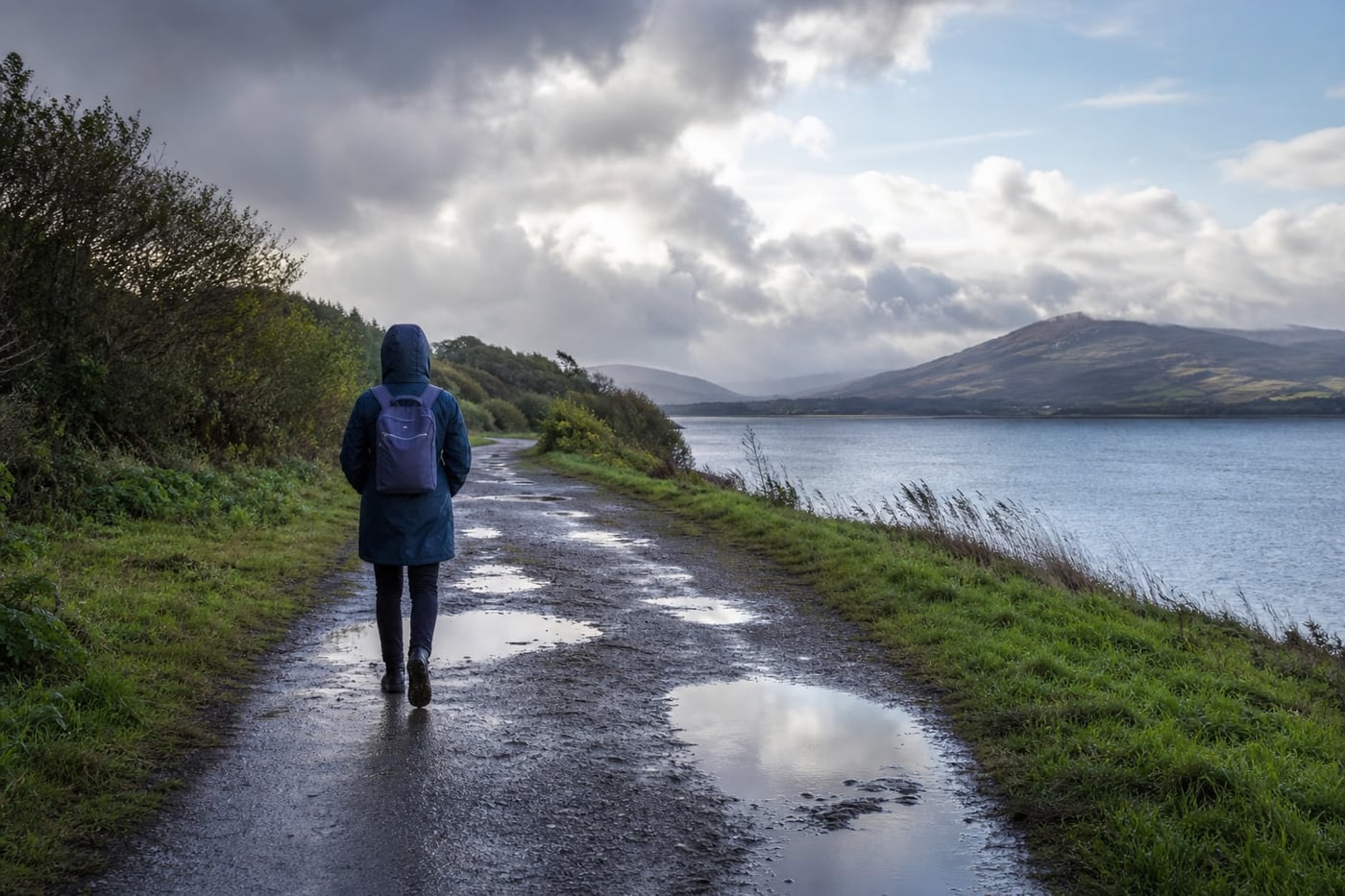 Person in a raincoat walking calmly along a quiet path after rainfall, carrying a small backpack beneath a clearing sky