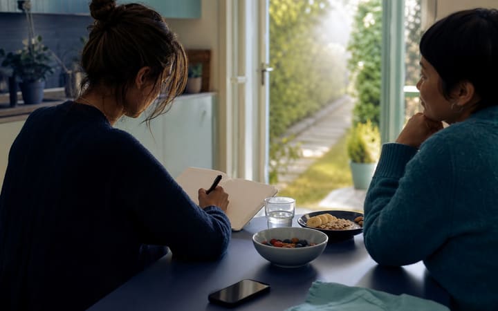 Person planning a sober day at a sunlit kitchen table with water, a notebook, and a supportive friend nearby