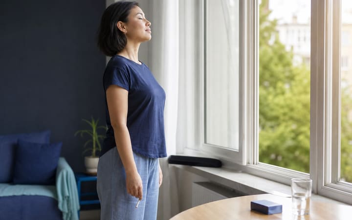 A person standing by an open window holding a broken unlit cigarette while taking a calm breath