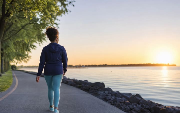 Person walking along a peaceful sunlit path beside trees and open water, symbolizing health, safety, and a new direction after alcohol.